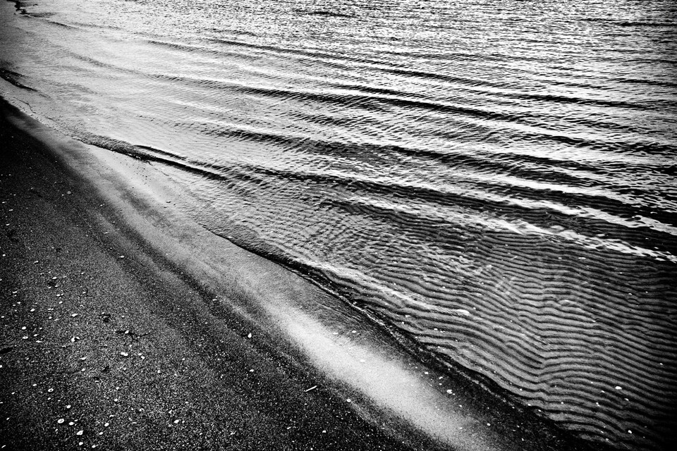 Krusat grunt vatten möter mörk sand i diagonal vattenlinje, närbild i svartvitt