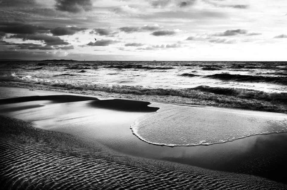 Vågskum sveper i båge över räfflad sand, brytande vågor under molnhimmel