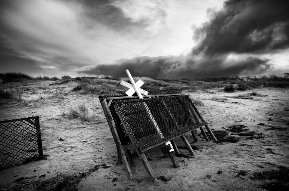 Gallerstaket med vitt kryss på sandstrand under hotande ovädersmoln, svartvitt