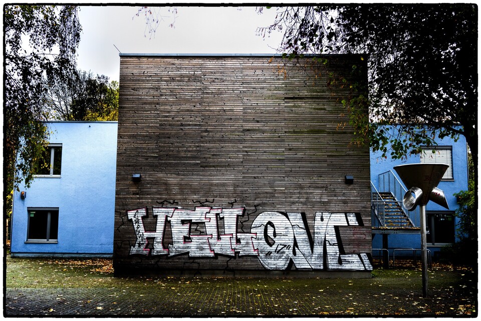 Graffiti på träfasad mellan blå byggnader, höstlöv på marken
