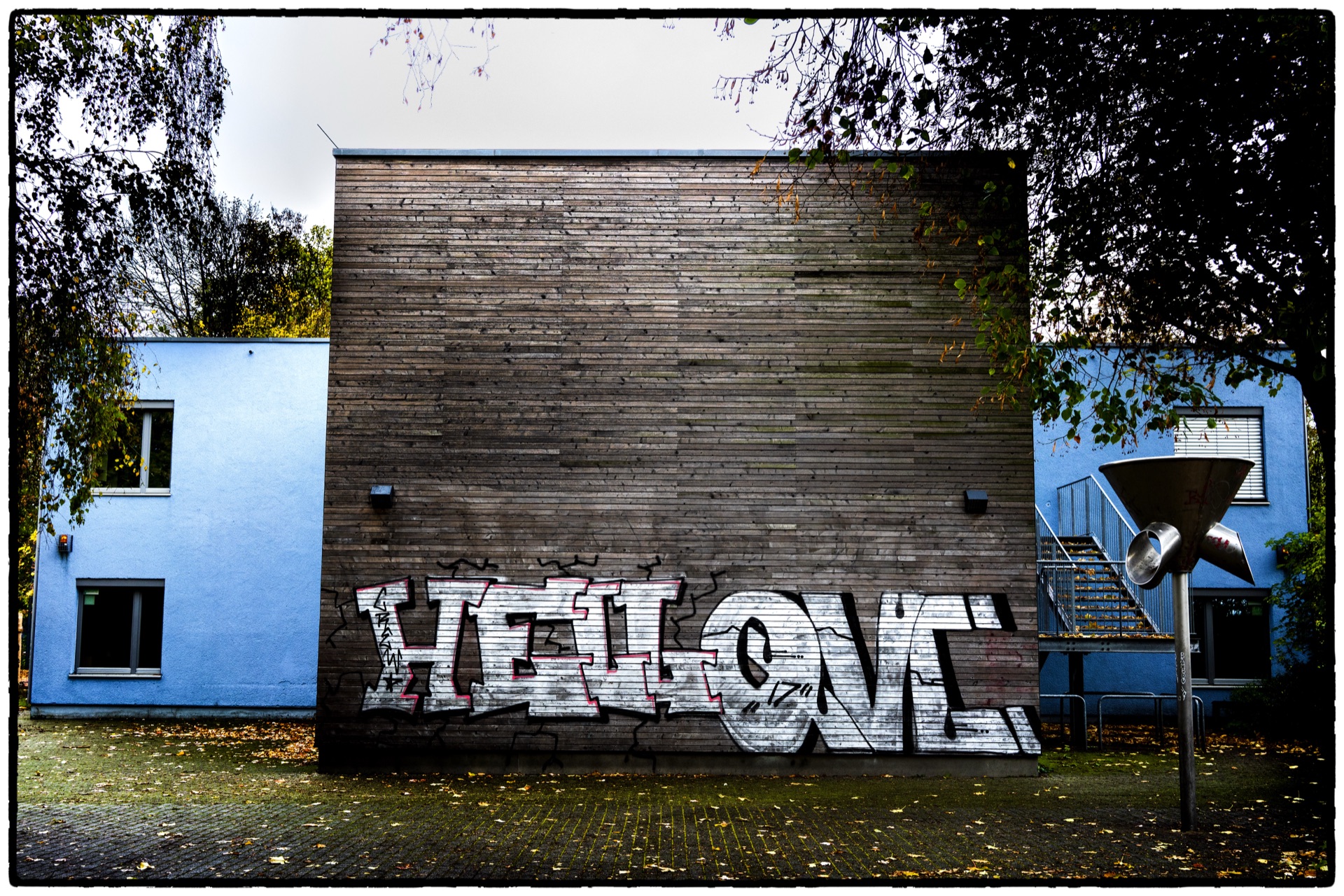Graffiti på träfasad mellan blå byggnader, höstlöv på marken