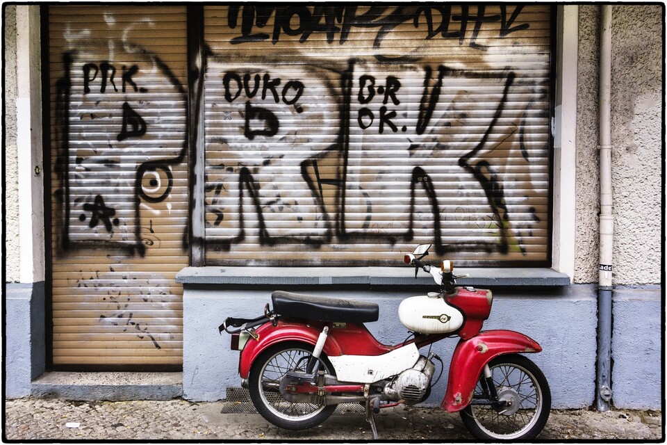 Röd veteranmoped parkerad framför neddragna jalusier täckta av graffiti