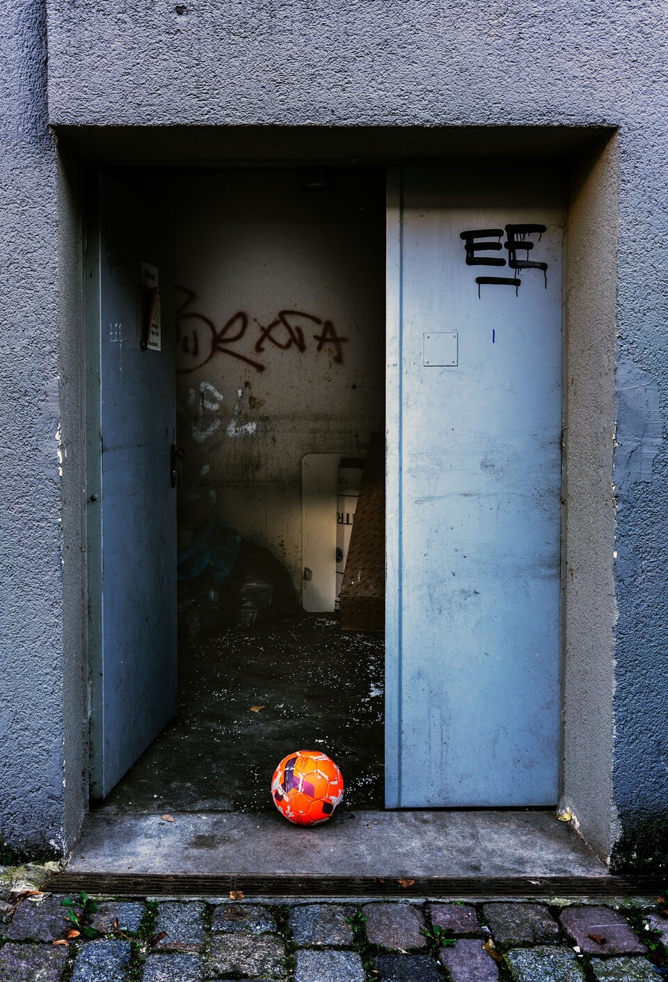 Orange fotboll i öppen dörröppning mot blågrå betongvägg