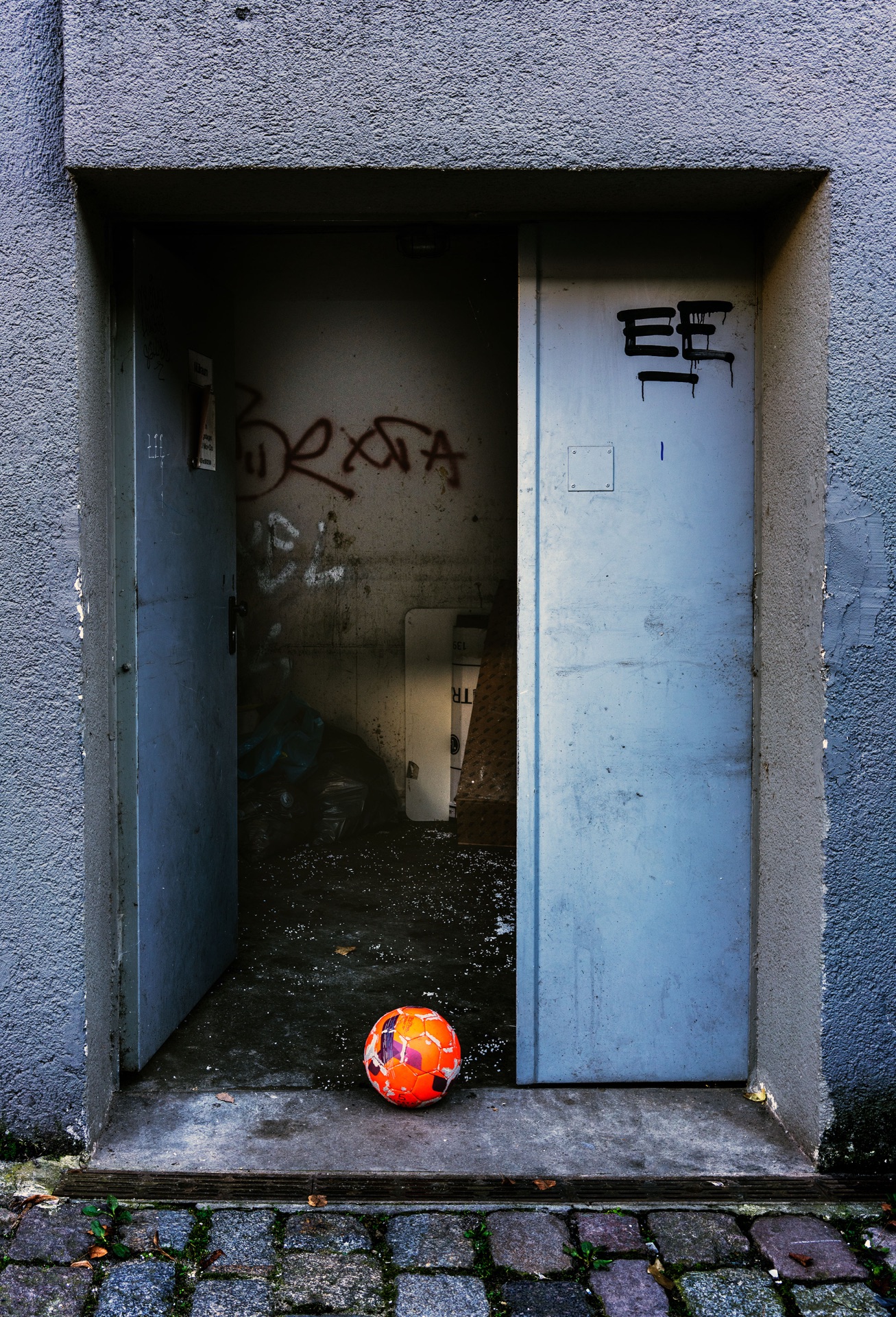 Orange fotboll i öppen dörröppning mot blågrå betongvägg