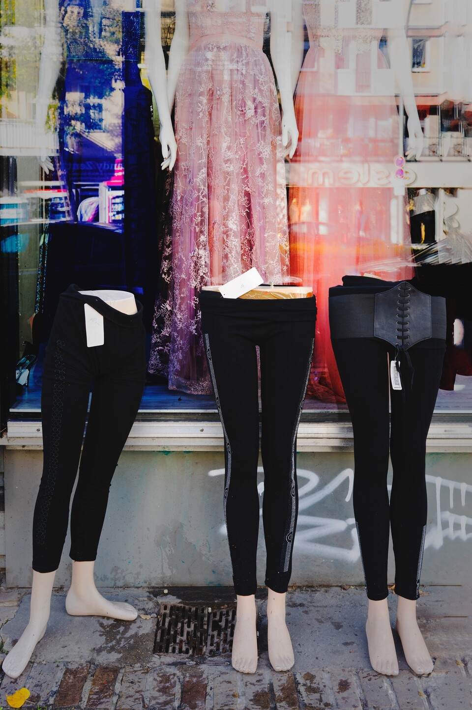 Tre skyltdocksben i svarta leggings på trottoaren framför fönster med rosa tyllklänning
