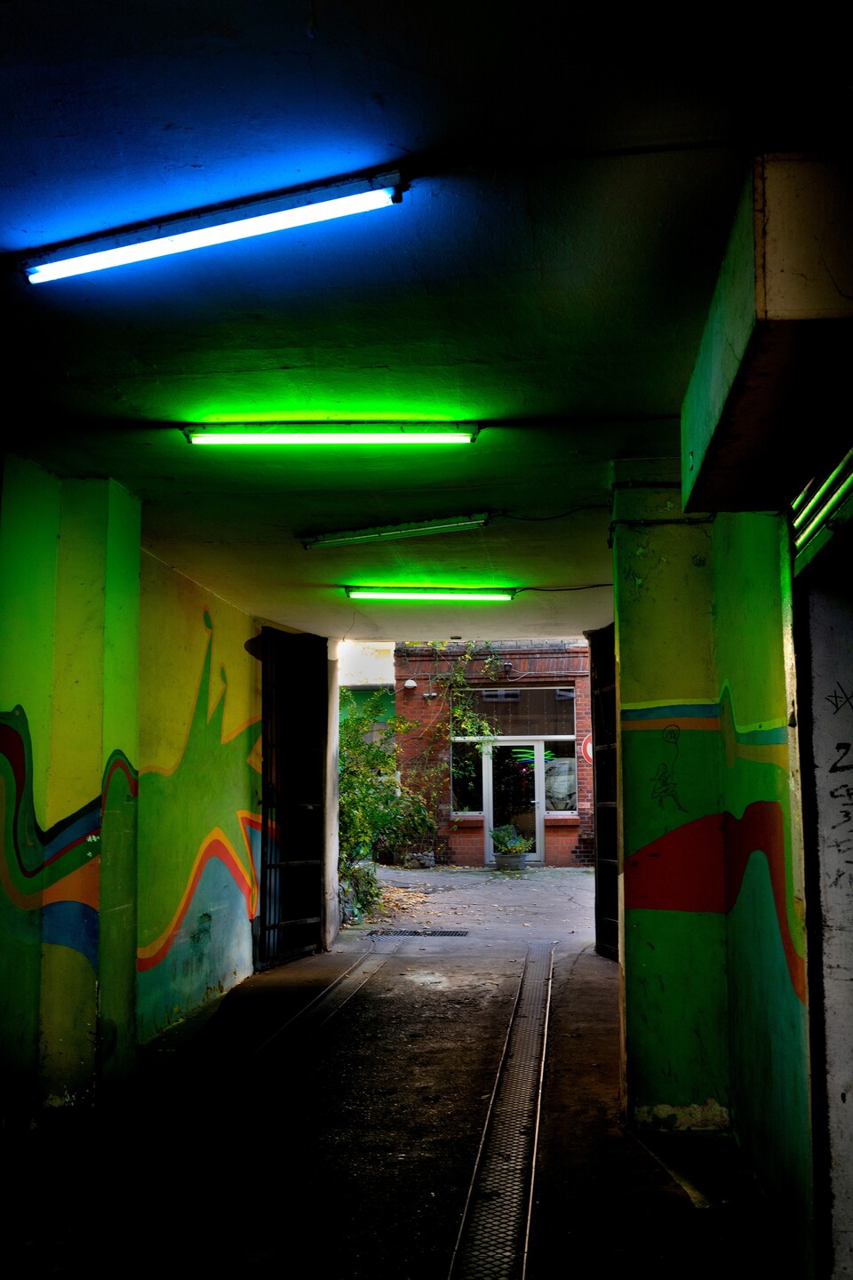 Lysrör i blått och grönt lyser upp graffitimålad passage mot grönskande innergård