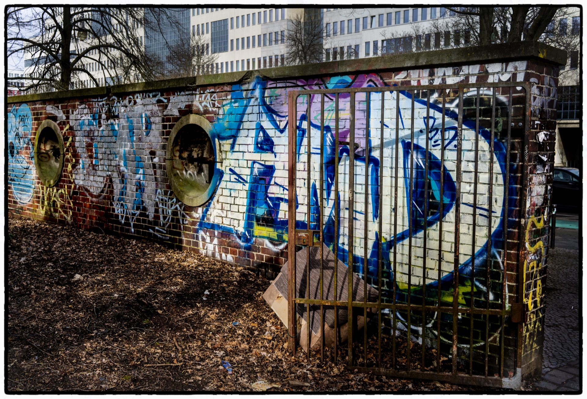 Blå graffiti på tegelvägg bakom metallgaller, höstlöv på marken