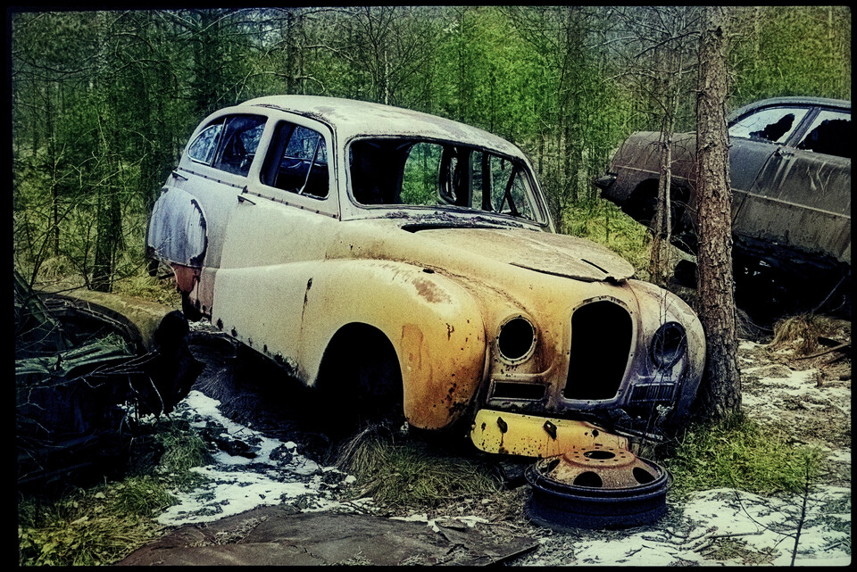 Gulvitt bilvrak utan hjul i skogsglänta, motorhuv och fälg i mossan intill