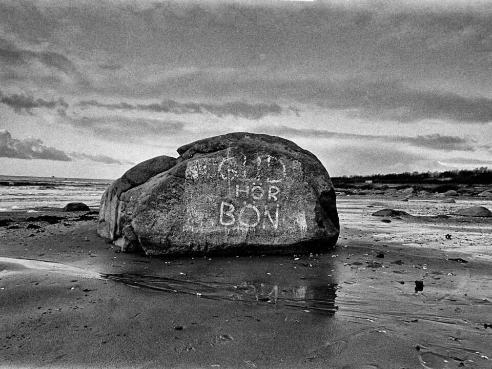 Stort stenblock med den målade inskriptionen Gud hör bön på våt strand, svartvitt