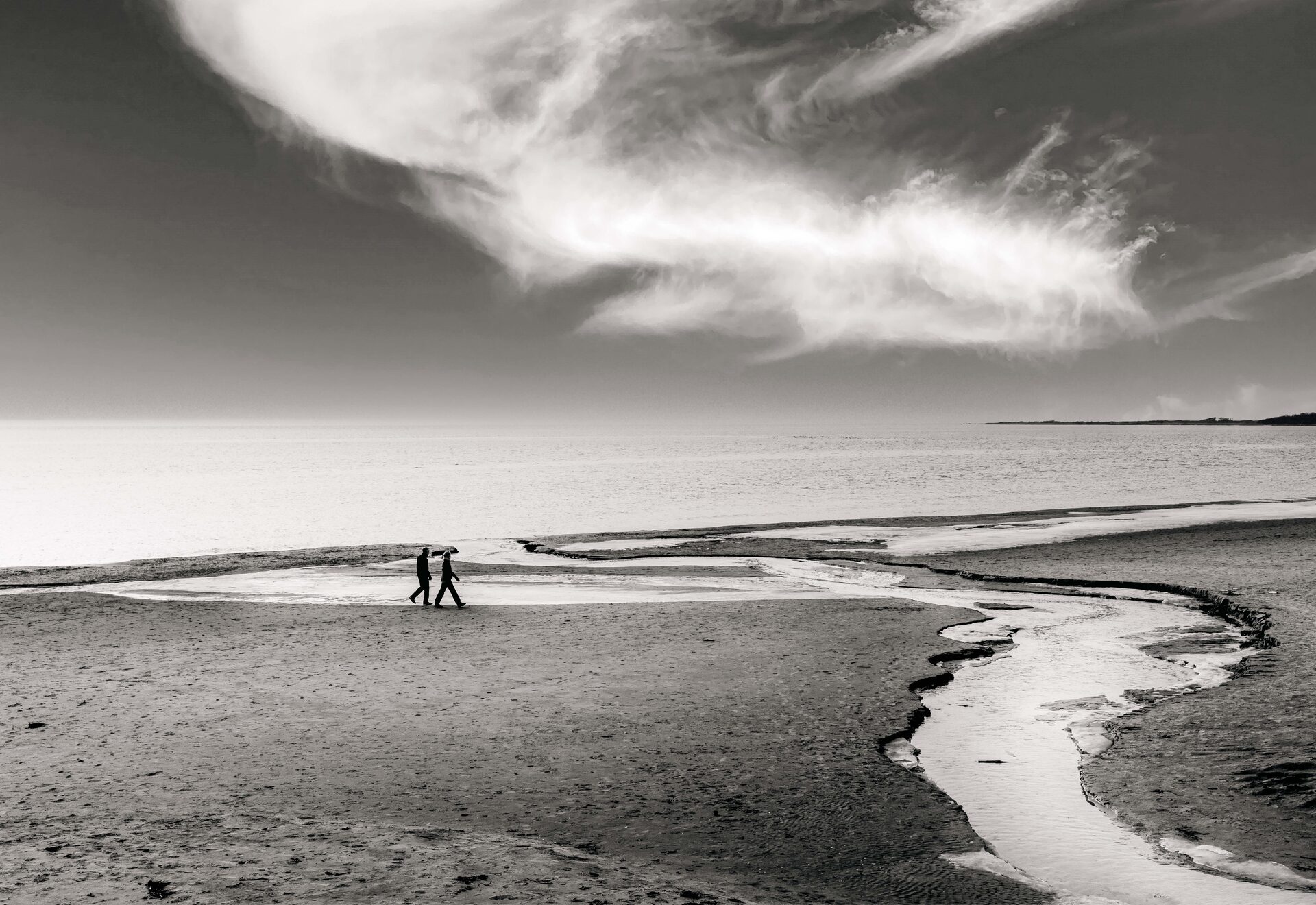Två figurer promenerar längs strandkant med vattenkanal och dramatisk himmel
