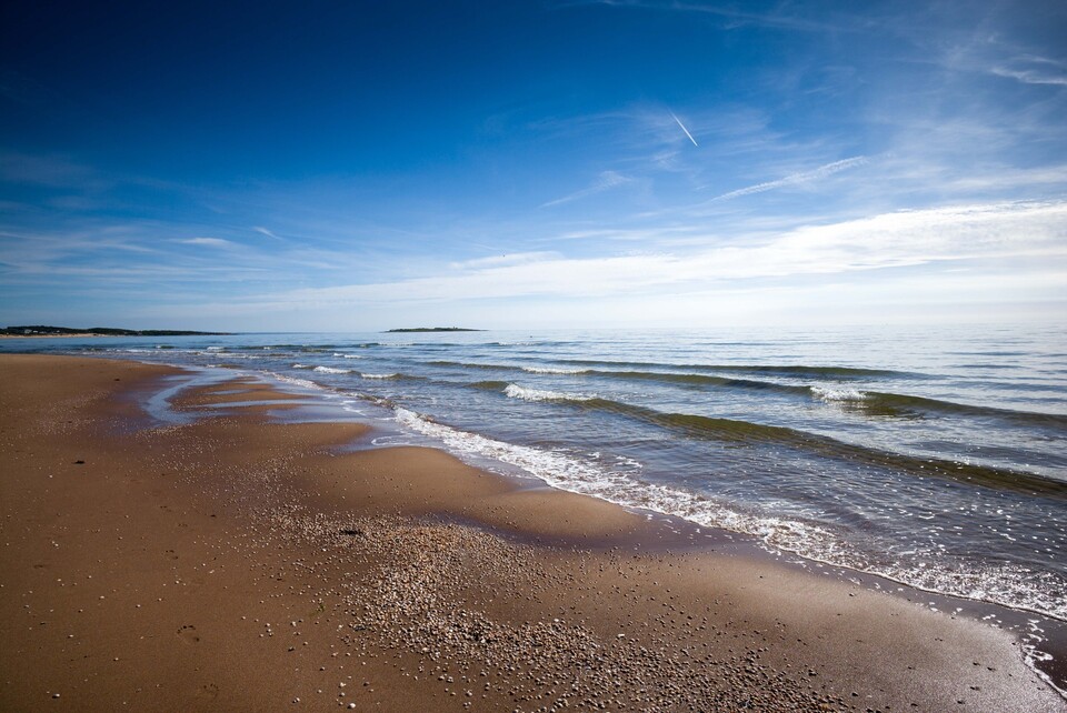V&aring;gor som rullar in mot grusig strandkant under klarbl&aring; sommarhimmel