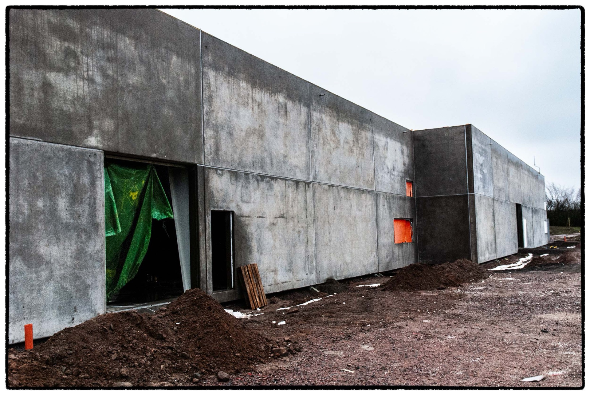 Rå betongbyggnad med gröna presenningar och lera i förgrunden