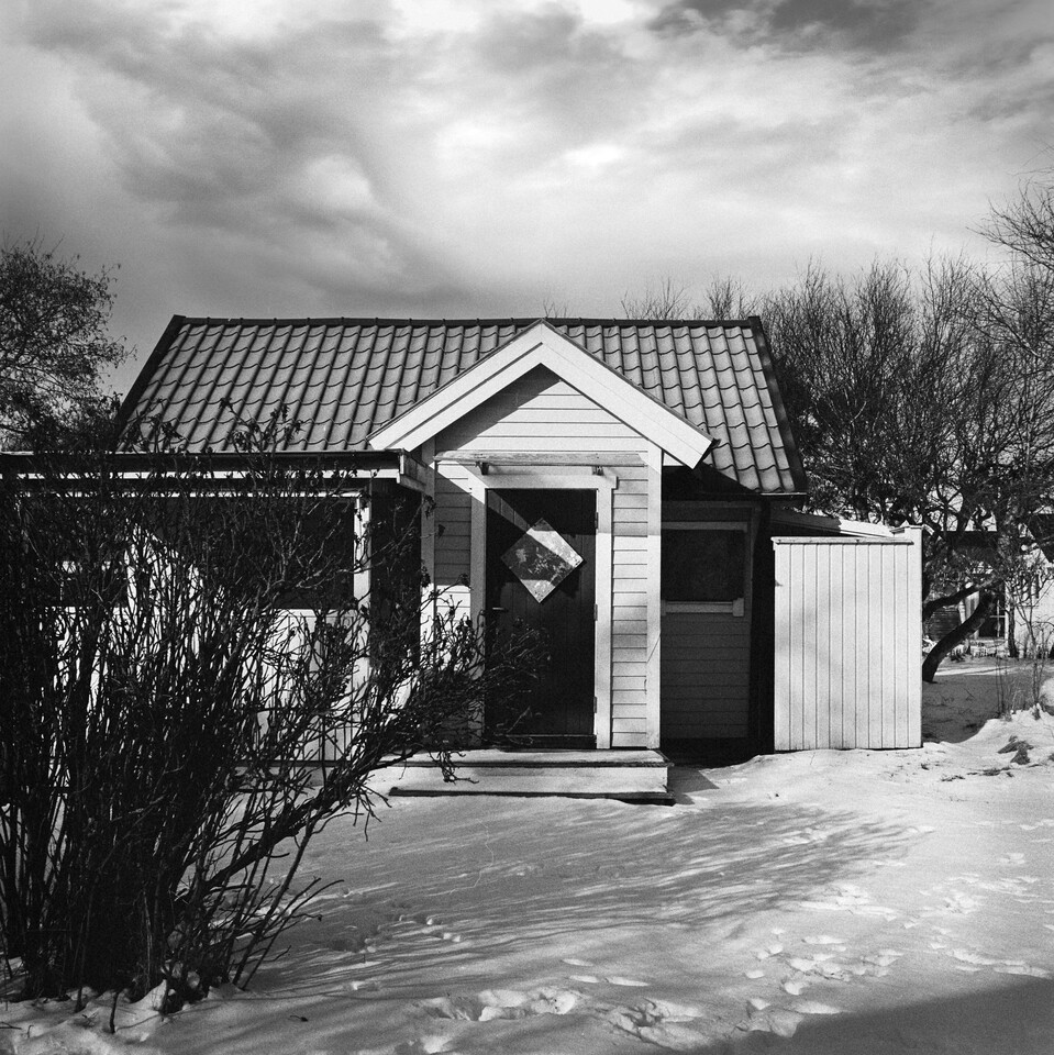 Liten stuga med tegeltak och farstukvist i sn&ouml;, avlövade buskar intill