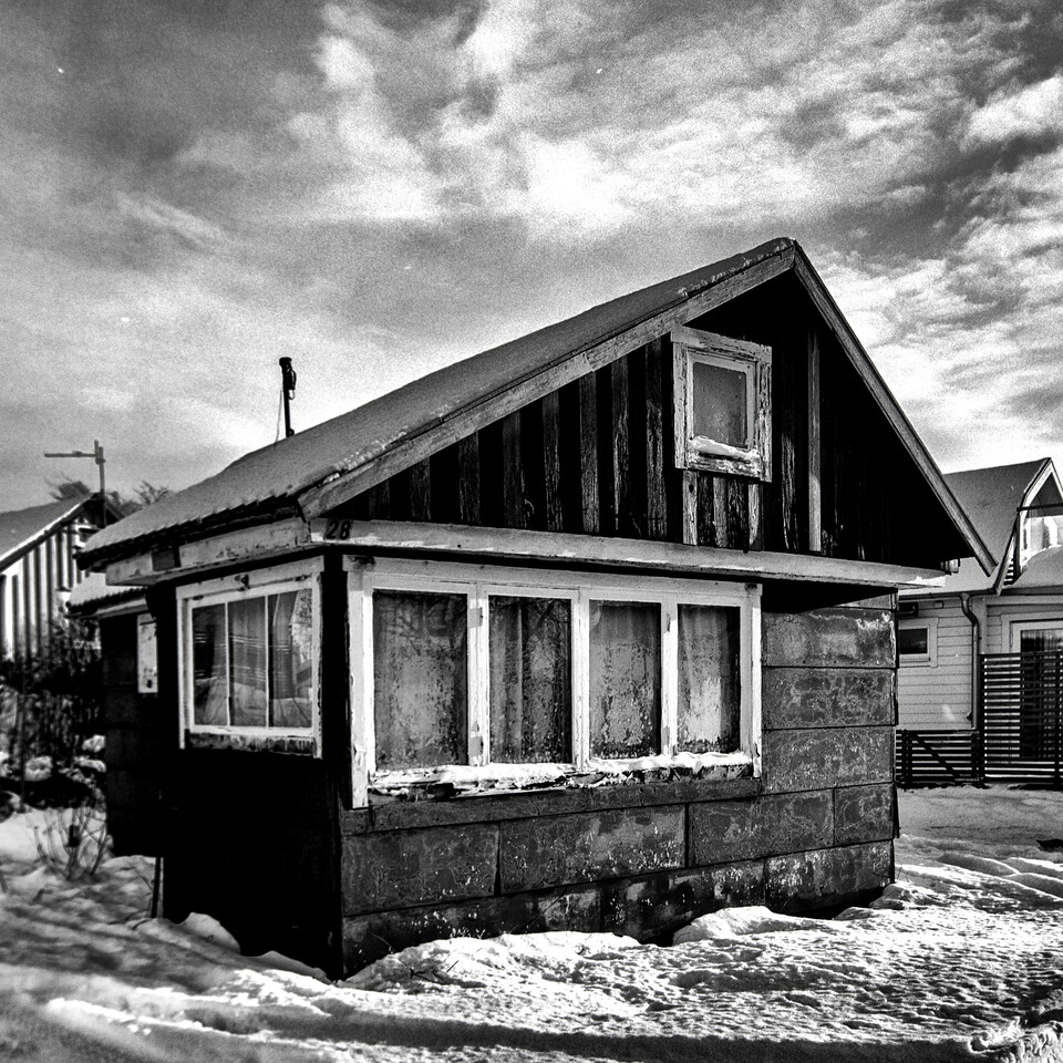 Vinklad strandstuga med igenspikade f&ouml;nster i sn&ouml;, grannhus bakom