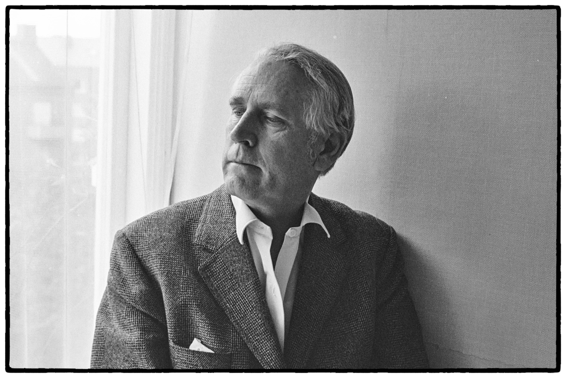 Äldre man i tweedkavaj blickar ut genom fönster, svartvitt