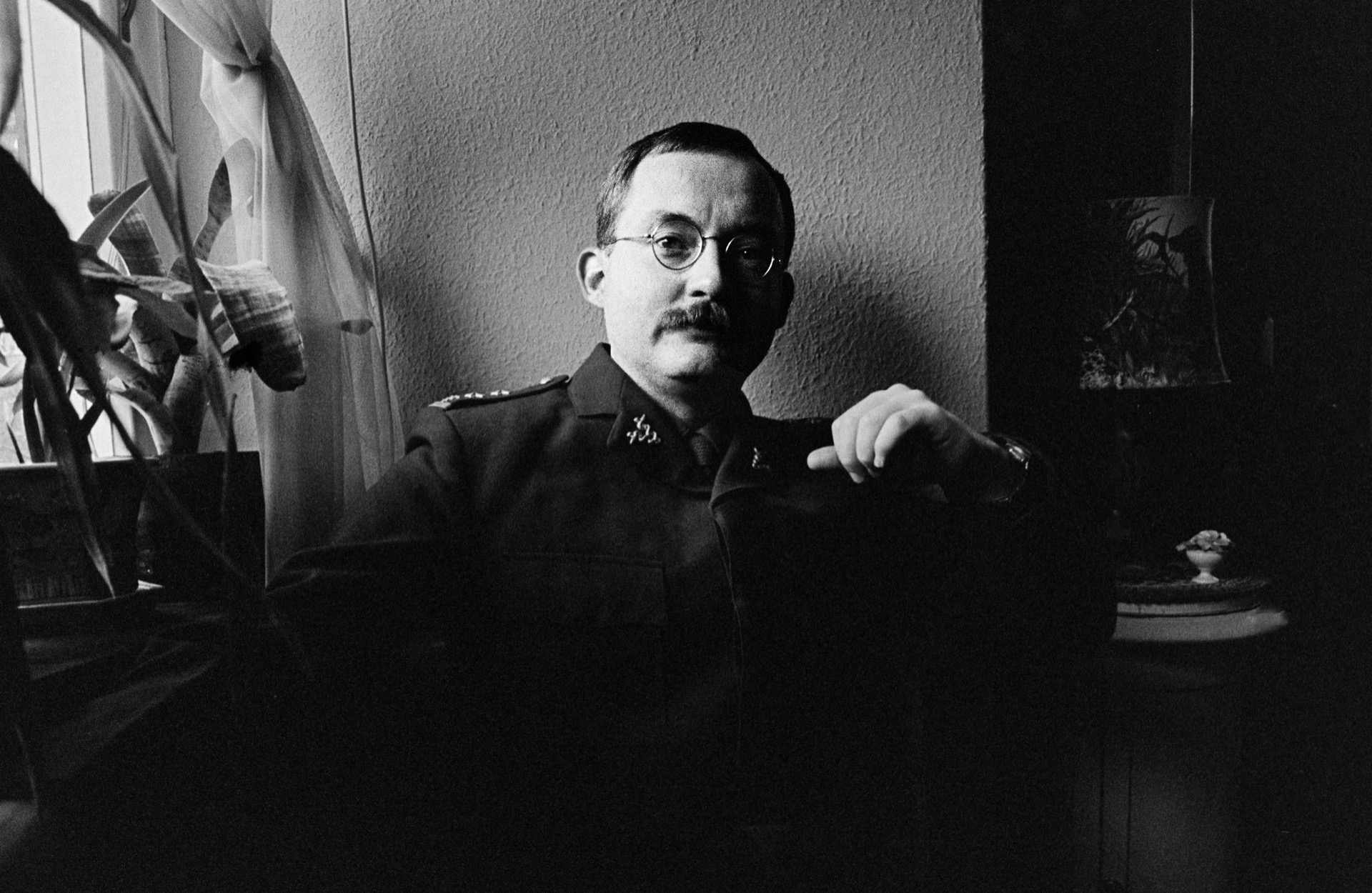 Man med mustasch och glasögon i uniform sitter vid fönster med krukväxt, svartvitt
