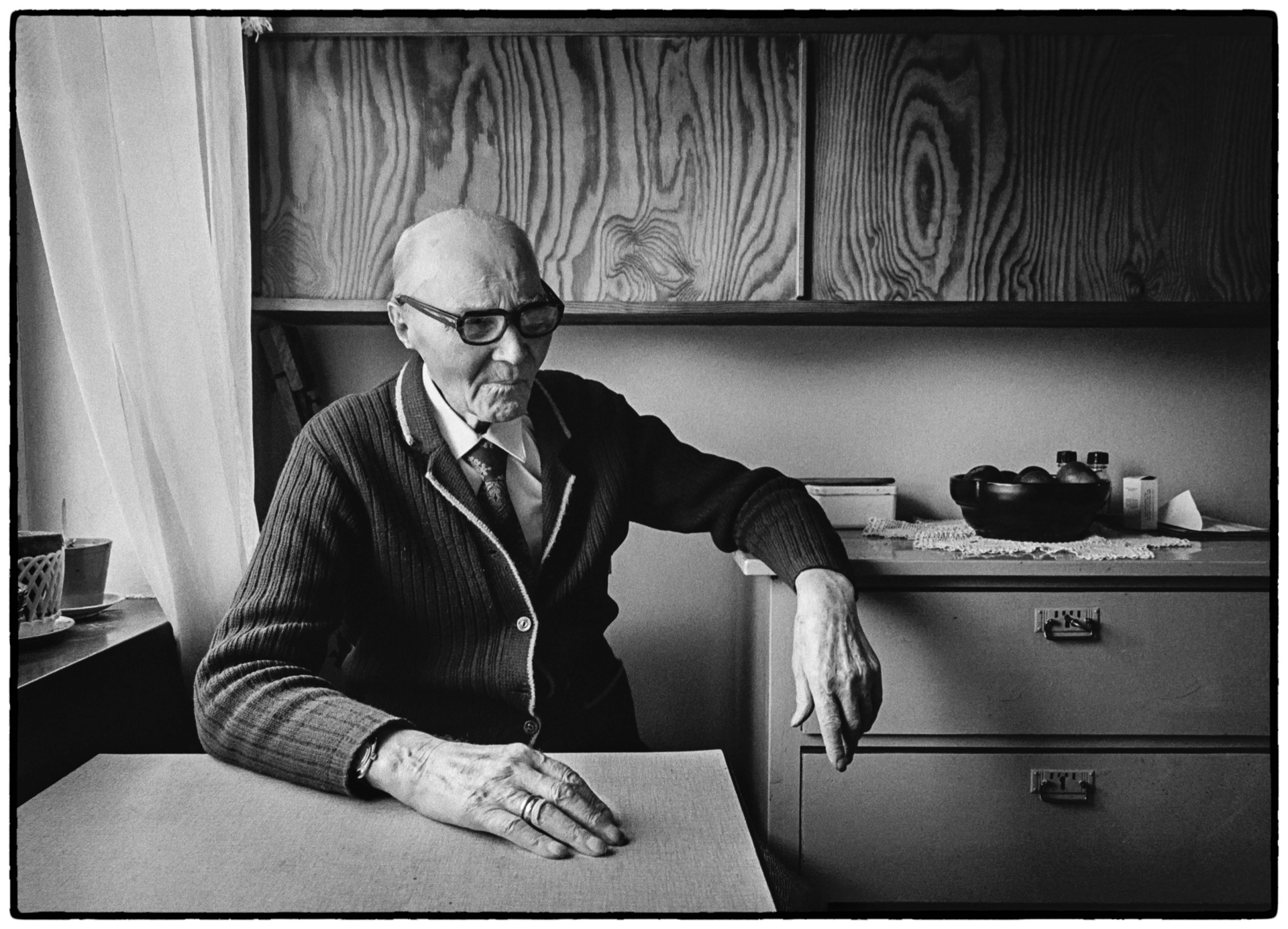 Äldre man med glasögon sitter vid köksbord, svartvitt