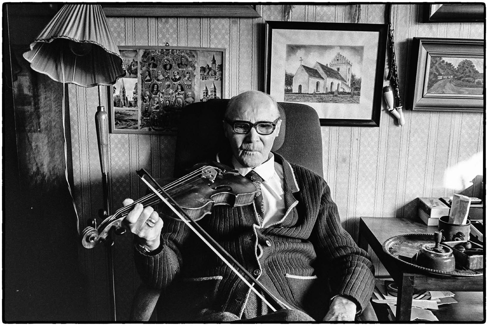 Äldre man med glasögon spelar fiol i sitt vardagsrum, svartvitt