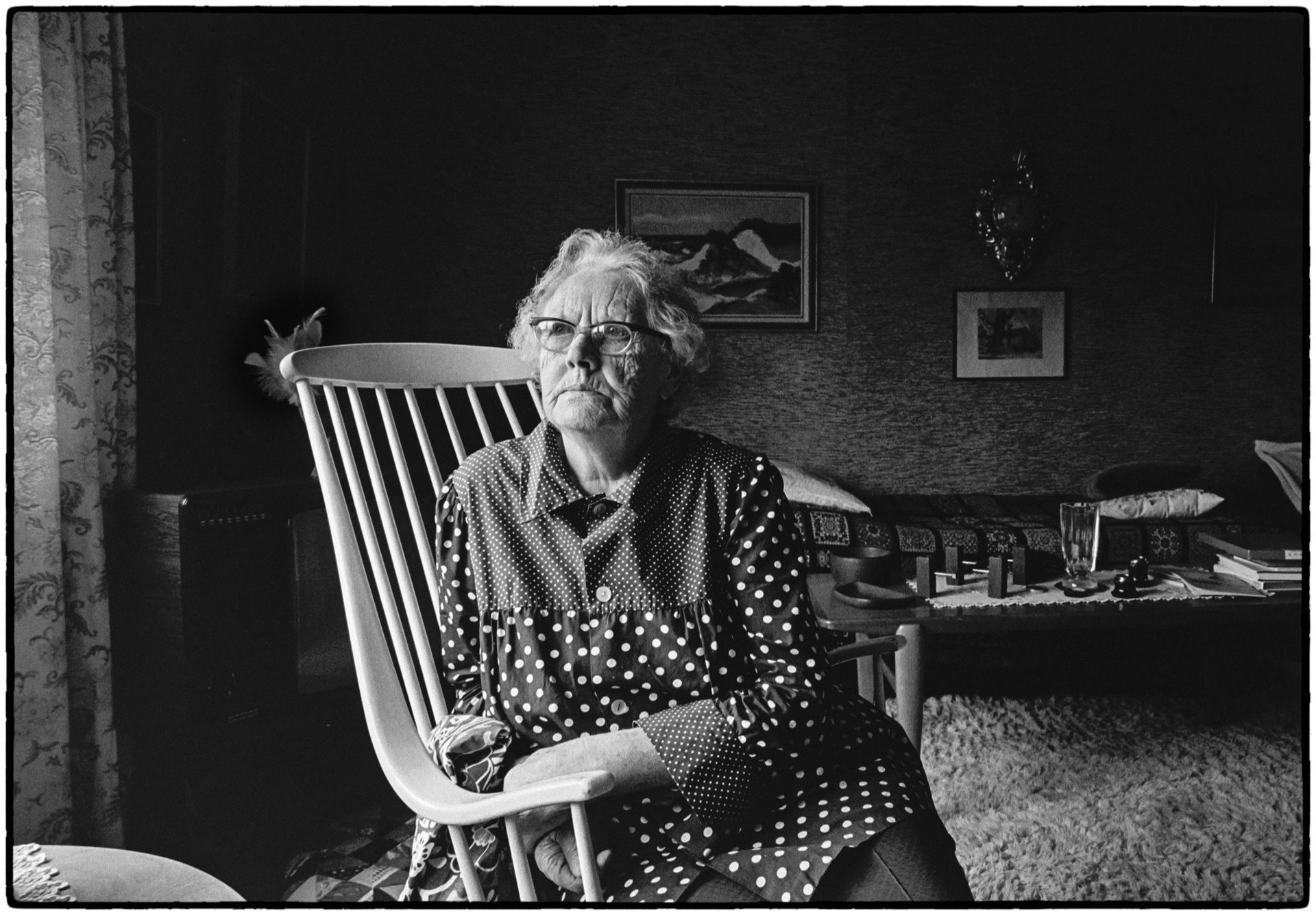 Äldre kvinna i prickig klänning sitter i gungstol, svartvitt