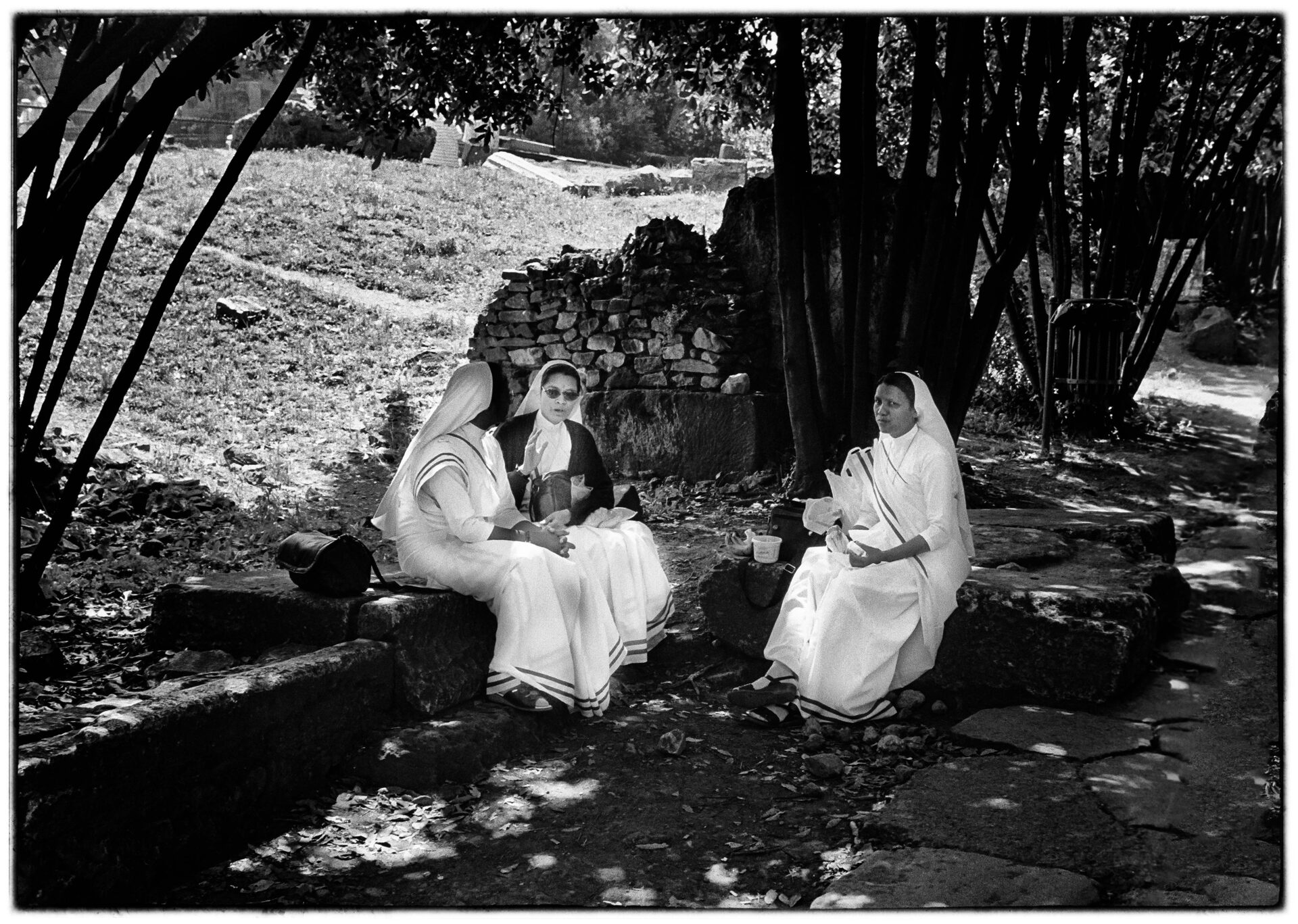 Tre nunnor vilar i skuggan under träd vid ruiner