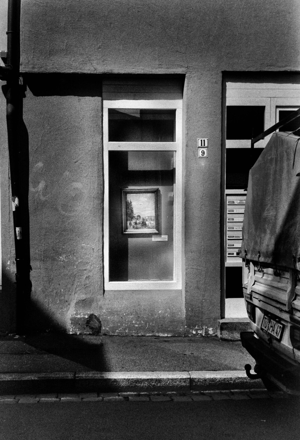 Inramad tavla i smalt skyltfönster, diagonal skugga över fasaden och kapelltäckt lastbil intill