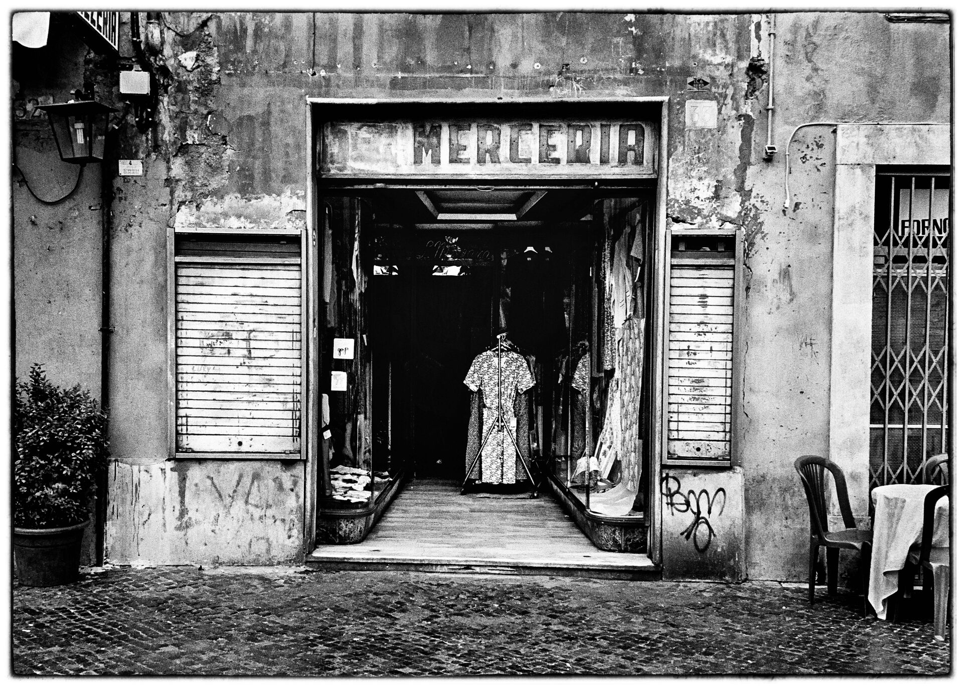 Merceria-skylt ovanför öppen butiksdörr med klänningar på vittrad fasad