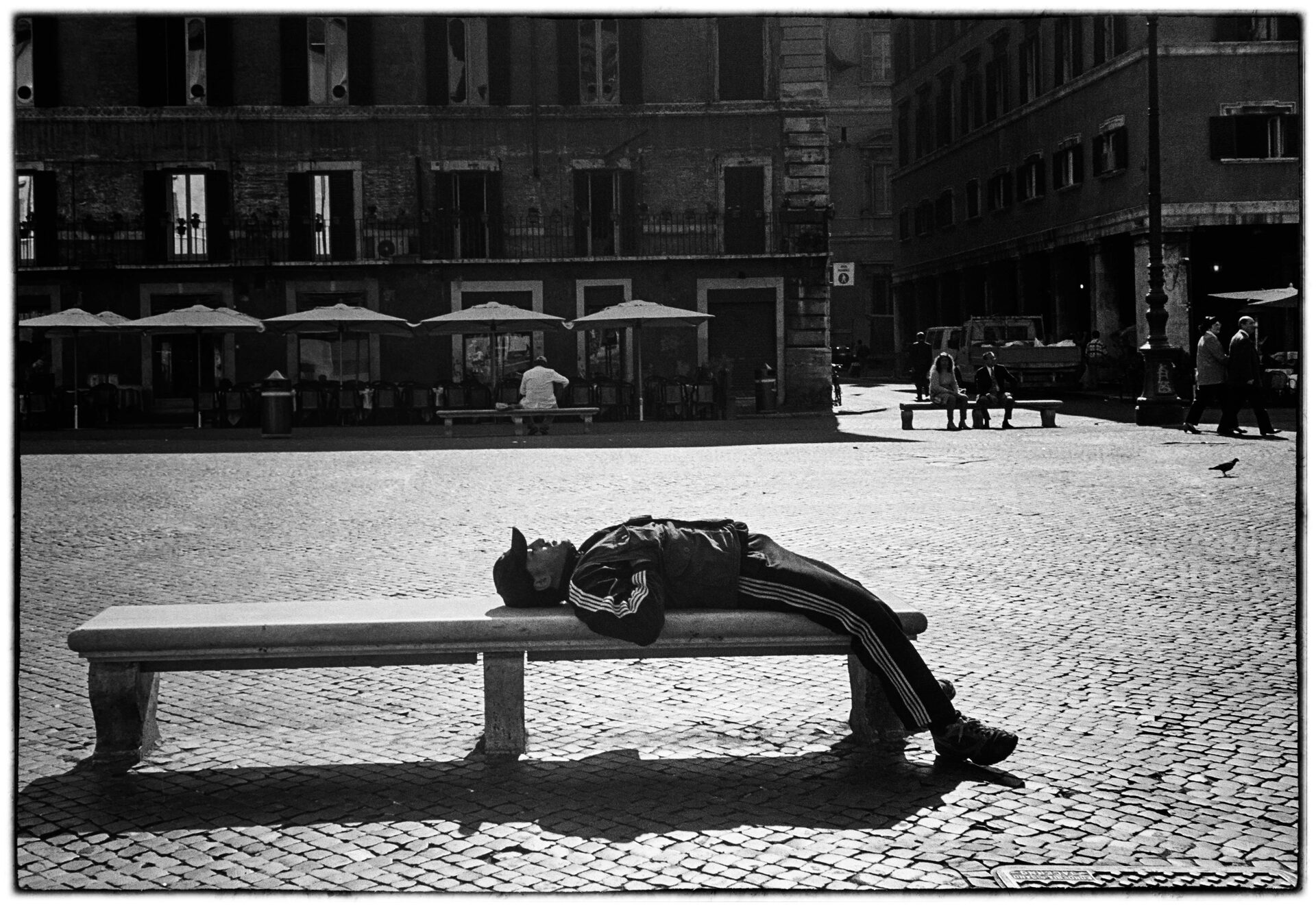 Man vilar på stenbänk på torg med fasader och parasoller i bakgrunden