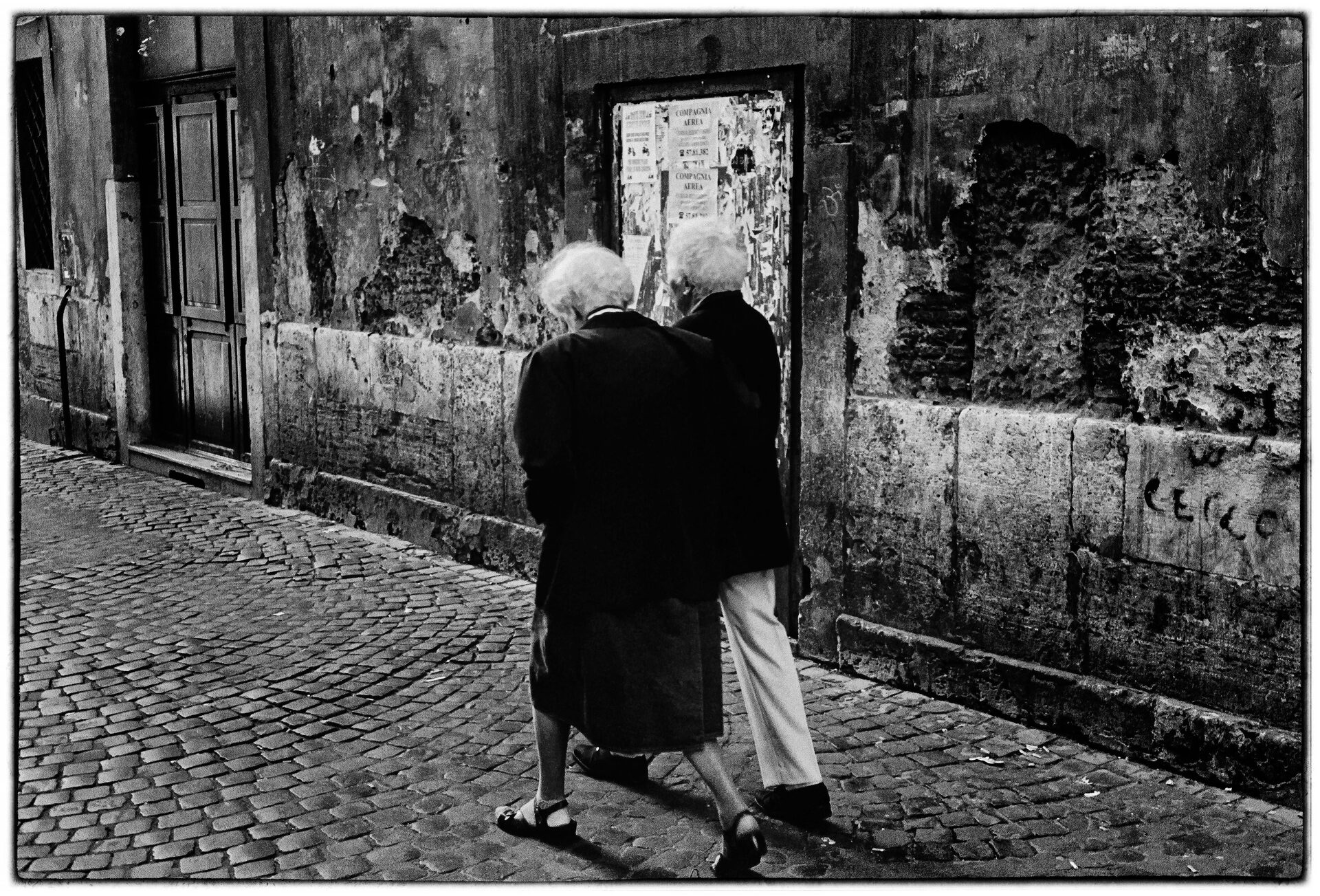 Äldre par arm i arm på kullerstensgata vid flagnad mur