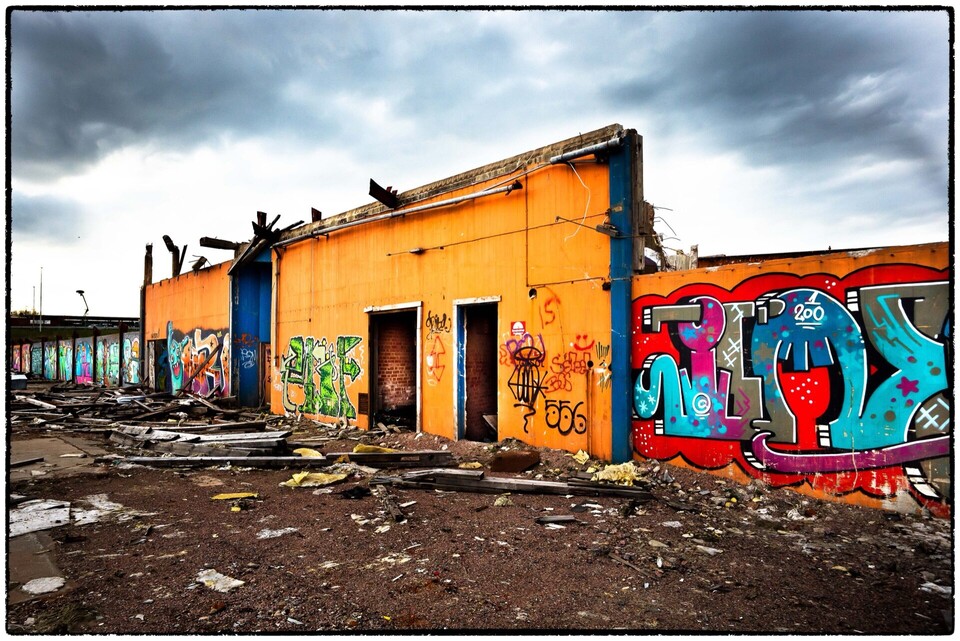 Orange fabriksruin utan tak med tomma dörröppningar, färgstark graffiti och bråte under mörka moln