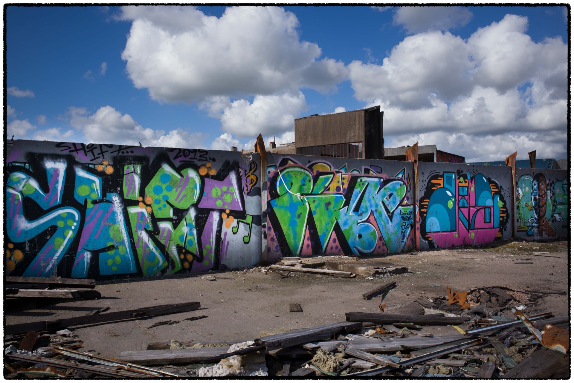 Blå och gröna graffitibokstäver på mur bland skrot